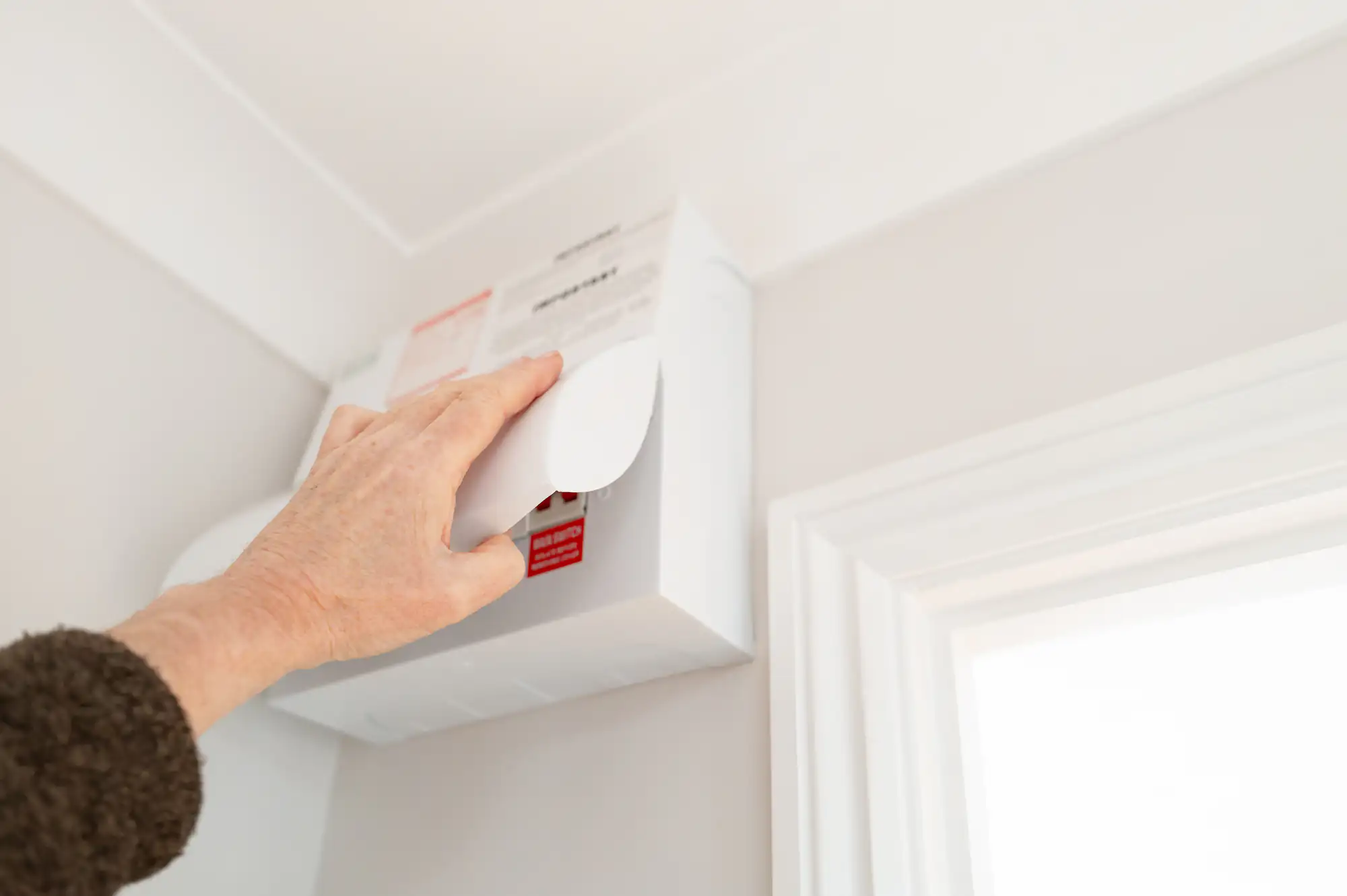 An electrician in Brooklyn, NY is opening the cover of an electrical fuse box mounted high on a light-colored wall near a window.
