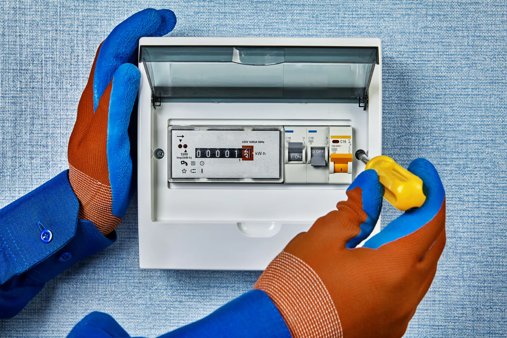 An electrician Brooklyn uses blue and orange protective gloves and a yellow screwdriver to adjust a circuit breaker switch inside an electrical meter box mounted on a textured blue wall in NY.