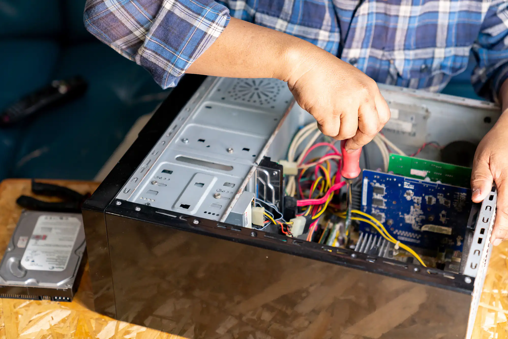 A person in a plaid shirt, resembling an electrician Brooklyn might trust, uses a screwdriver to work inside an open desktop computer case. Various cables and components are visible, with a hard drive and pliers on the table nearby in NY.