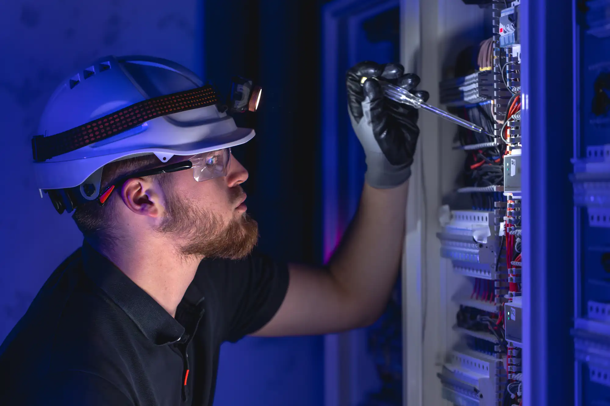 An electrician Brooklyn professional, wearing safety gear like a helmet, goggles, gloves, and a headlamp, uses a screwdriver to work on electrical panels filled with wires in a dimly lit NY environment.