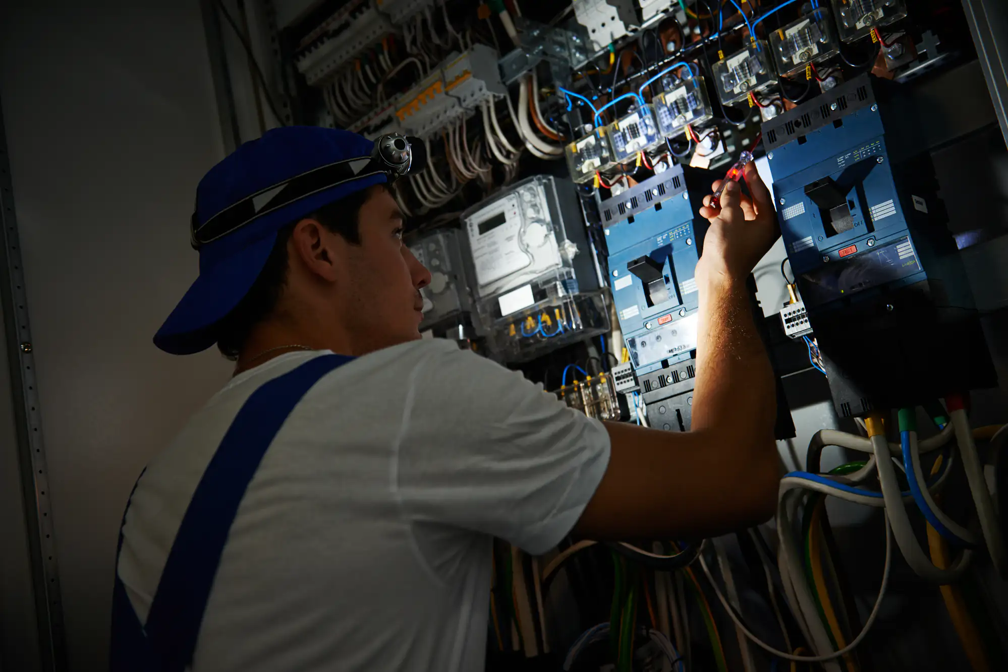 An electrician in Brooklyn, NY, wearing a blue cap, headlamp, and white shirt, adjusts electrical components inside a control panel filled with wires and switches in a technical workspace.