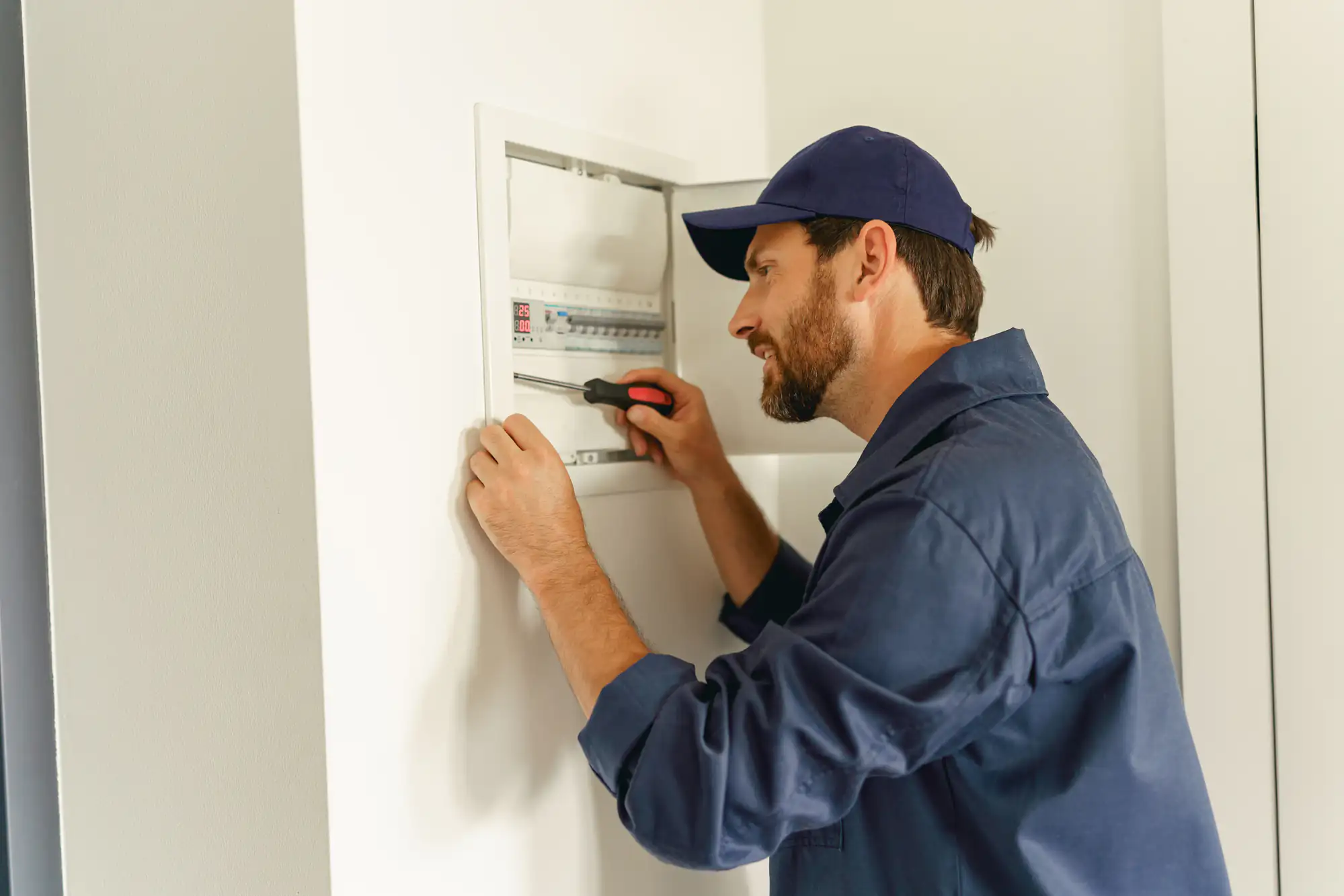 A man in a blue cap and work uniform, an electrician Brooklyn trusts, uses a screwdriver to repair or inspect an electrical panel on a white wall in NY.