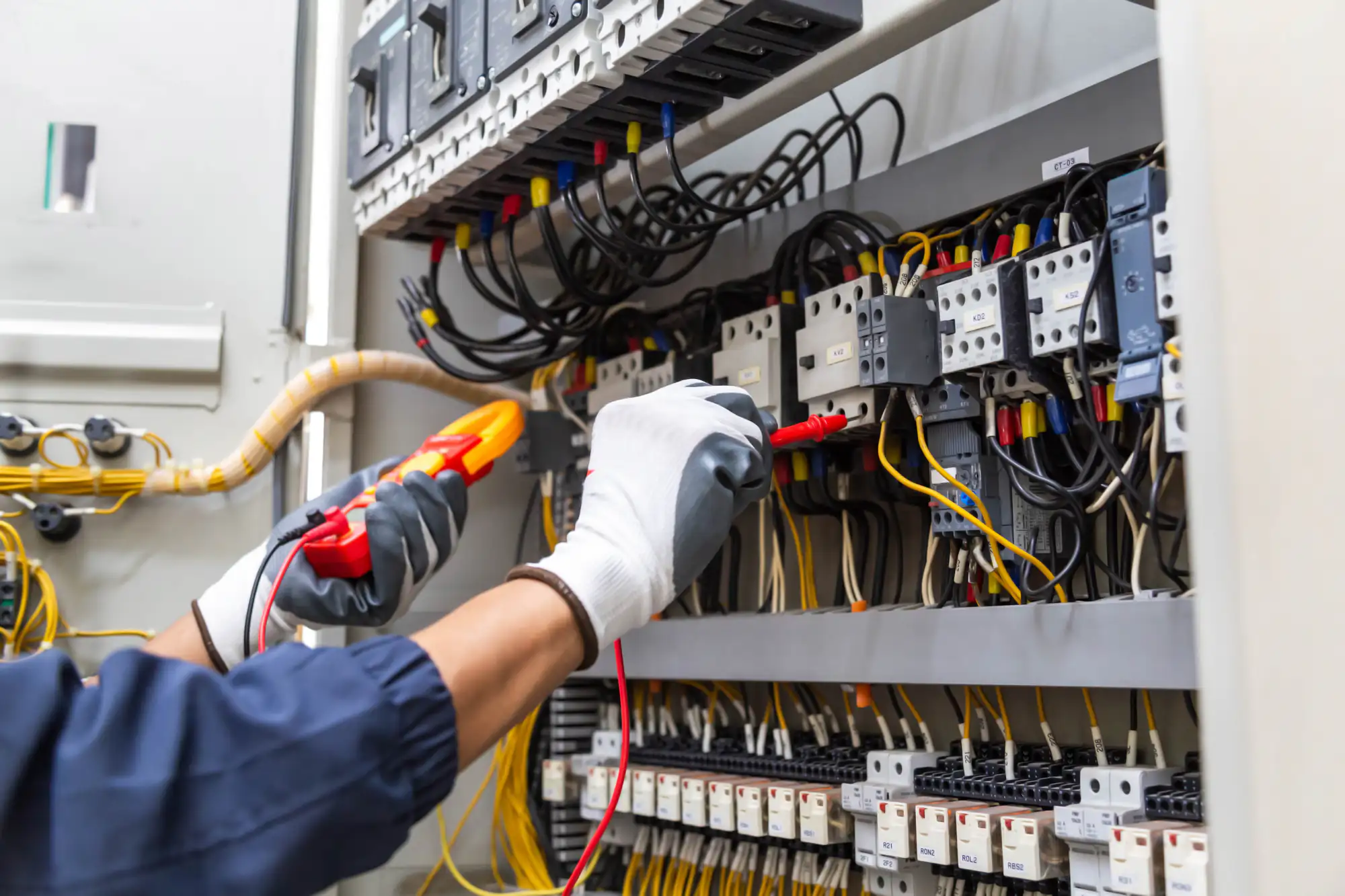 An electrician Brooklyn wearing white gloves uses a digital multimeter to test electrical circuits inside an open control panel filled with wires and switches in NY.