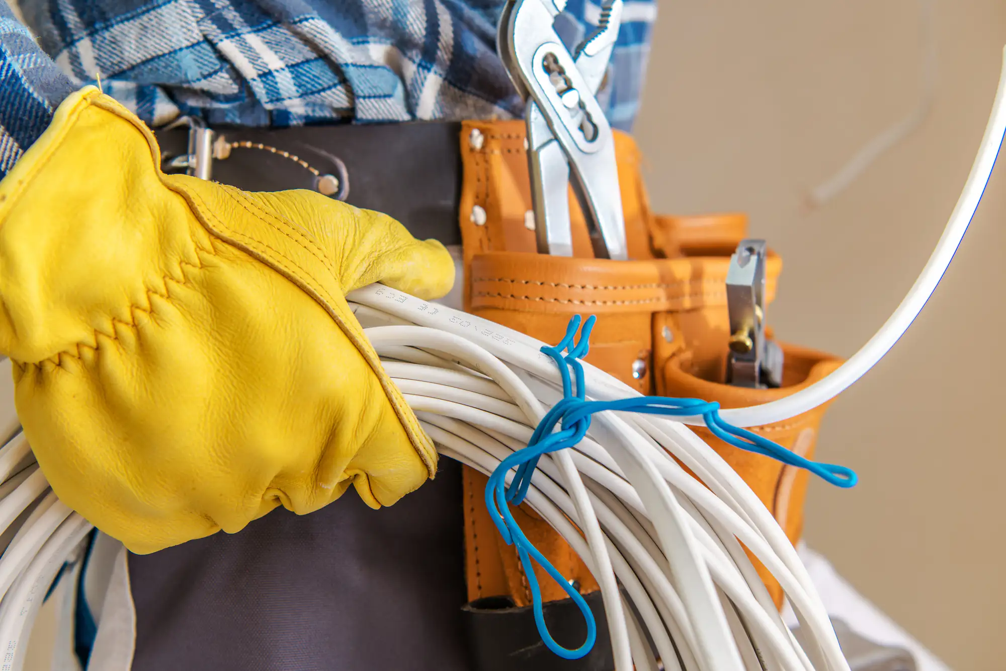 An electrician in Brooklyn, NY, wearing yellow work gloves and a tool belt, holds a coiled bundle of white electrical cables. Various tools are visible in leather pouches, and the individual is dressed in a plaid shirt.