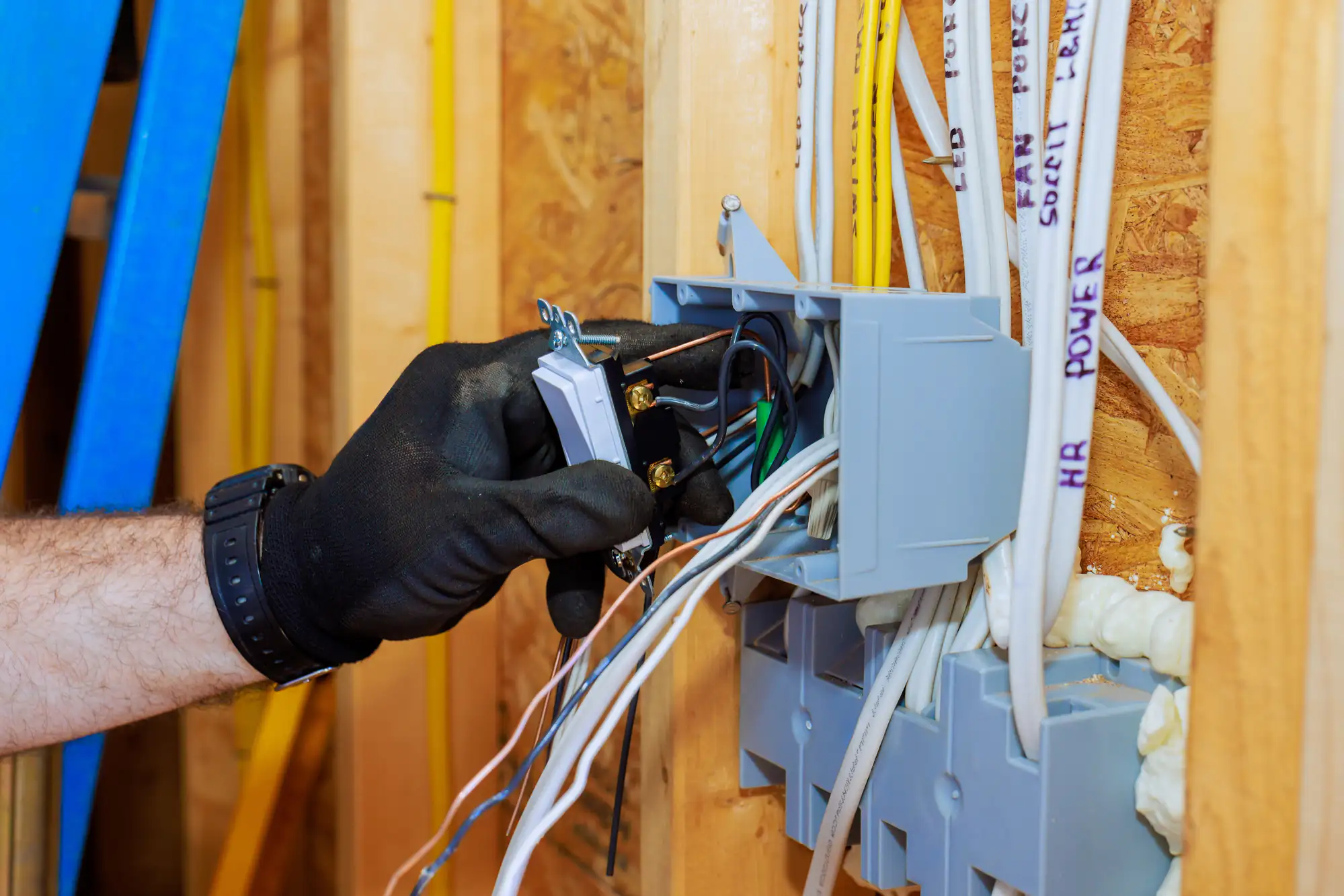 An electrician Brooklyn wearing a black glove installs electrical wiring and a light switch in a wall outlet box of a building under construction in NY, with exposed wooden framing and cables visible.