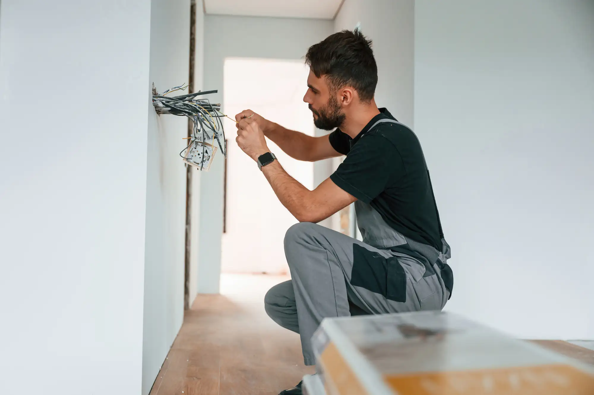 An electrician Brooklyn in work overalls kneels by a wall, working with exposed electrical wires in a partially finished NY room with wooden floors and white walls.
