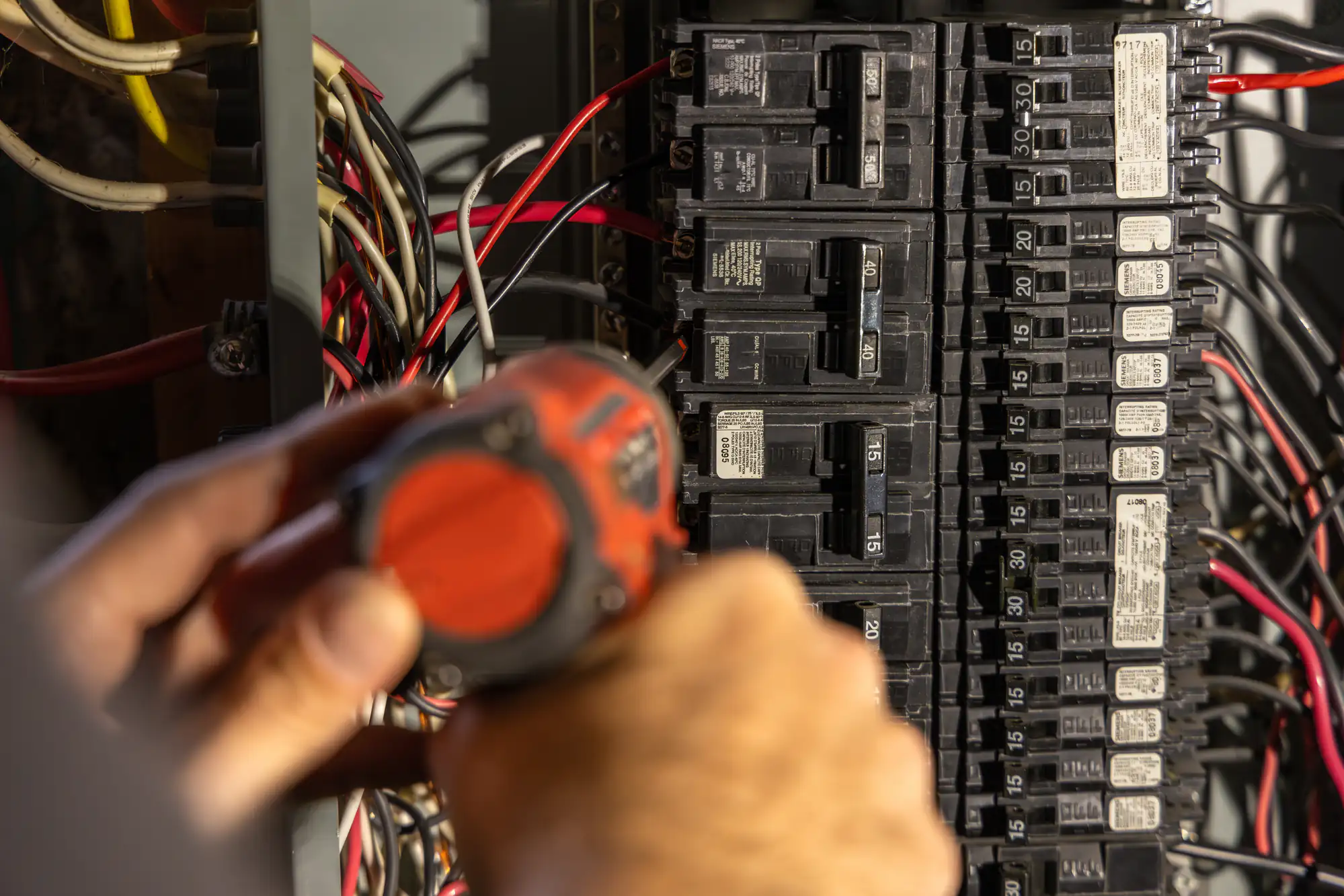 A skilled electrician Brooklyn uses an orange cordless drill to work on an electrical circuit breaker panel with multiple wires and switches in NY.