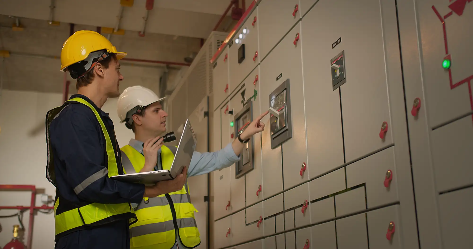 Two engineers wearing safety helmets and reflective vests stand in front of control panels&mdash;similar to an electrician in Brooklyn, NY&mdash;one holds a laptop while the other points at a screen, discussing or monitoring equipment in an industrial facility.