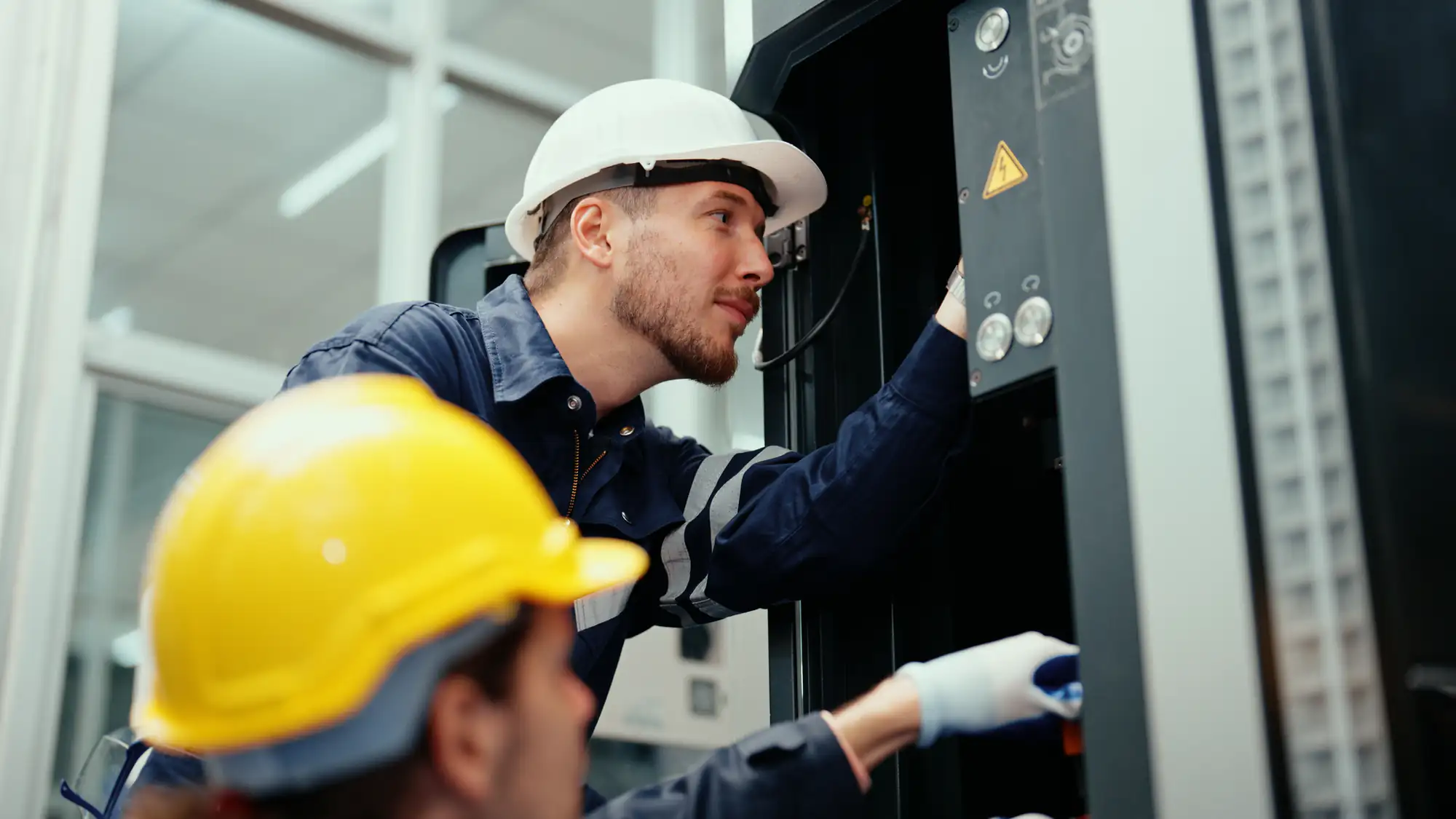 Two workers wearing safety helmets and gloves inspect and operate electrical equipment inside an industrial setting. The focus is on one electrician Brooklyn adjusting controls on a large panel in NY.