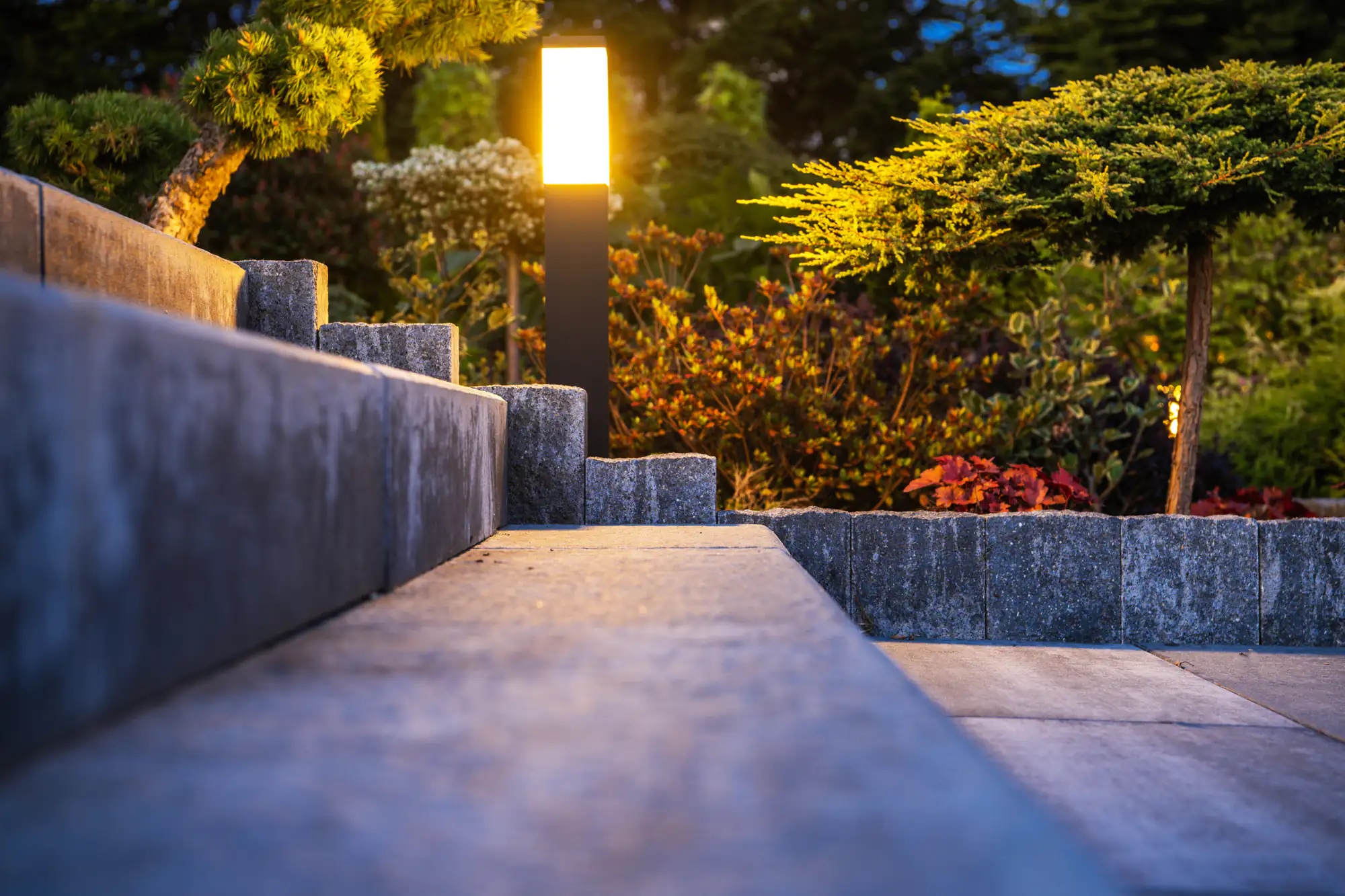 A modern NY garden scene at dusk, featuring stone steps, a glowing outdoor lamp installed by an electrician Brooklyn trusts, and surrounding greenery with neatly trimmed shrubs and colorful plants.