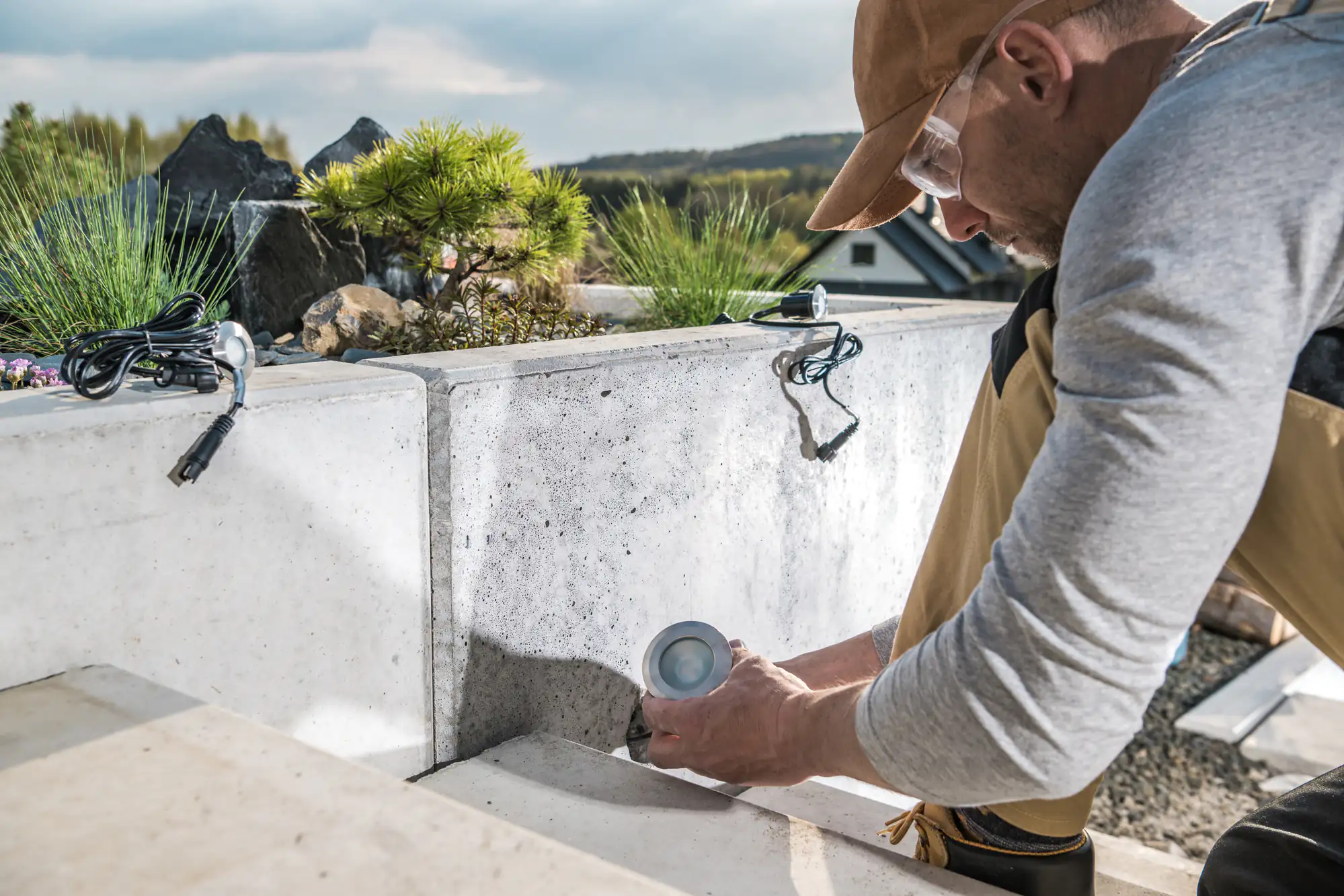 A person wearing safety glasses and a cap installs an outdoor light fixture into a concrete garden wall, with wiring and greenery in the background&mdash;a scene typical for an electrician Brooklyn, NY might call on for expert installations.