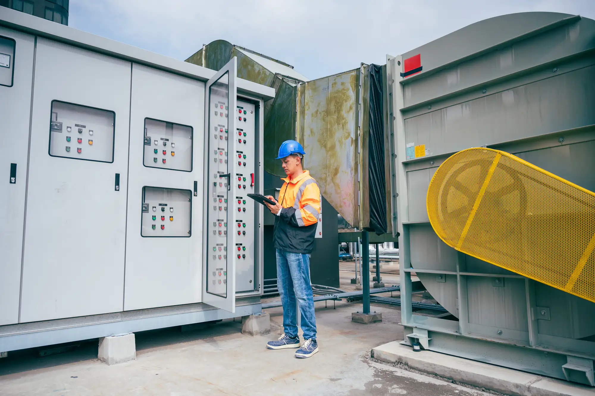 An electrician in Brooklyn, NY wearing a blue hard hat and safety vest inspects industrial electrical control panels outdoors, holding a clipboard near large machinery on a concrete surface.