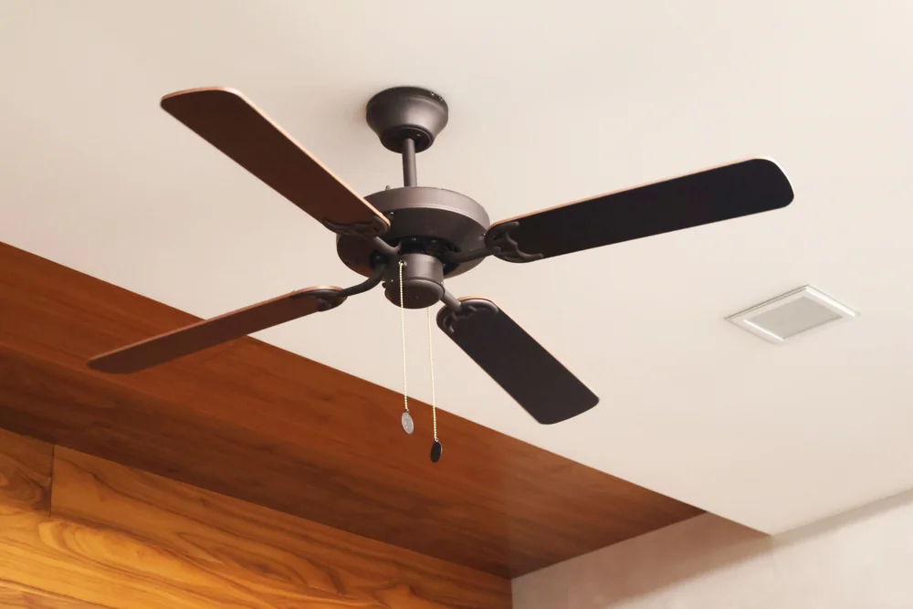A black ceiling fan with four blades and pull chains is mounted on a white ceiling with wooden panel accents.