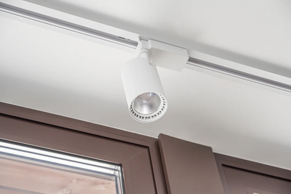 A white ceiling-mounted track light fixture is installed above a window with a brown frame, illuminating the area below.