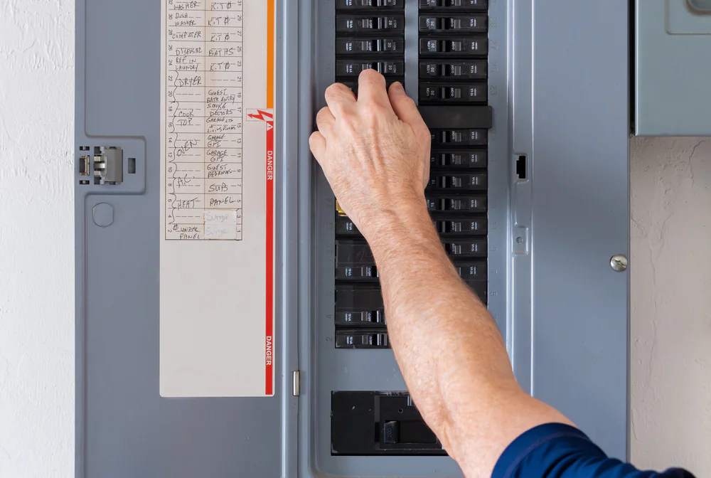 A person&rsquo;s hand is flipping a switch inside a gray electrical breaker panel mounted on a wall, with a list of circuit labels visible on the inside of the panel door.