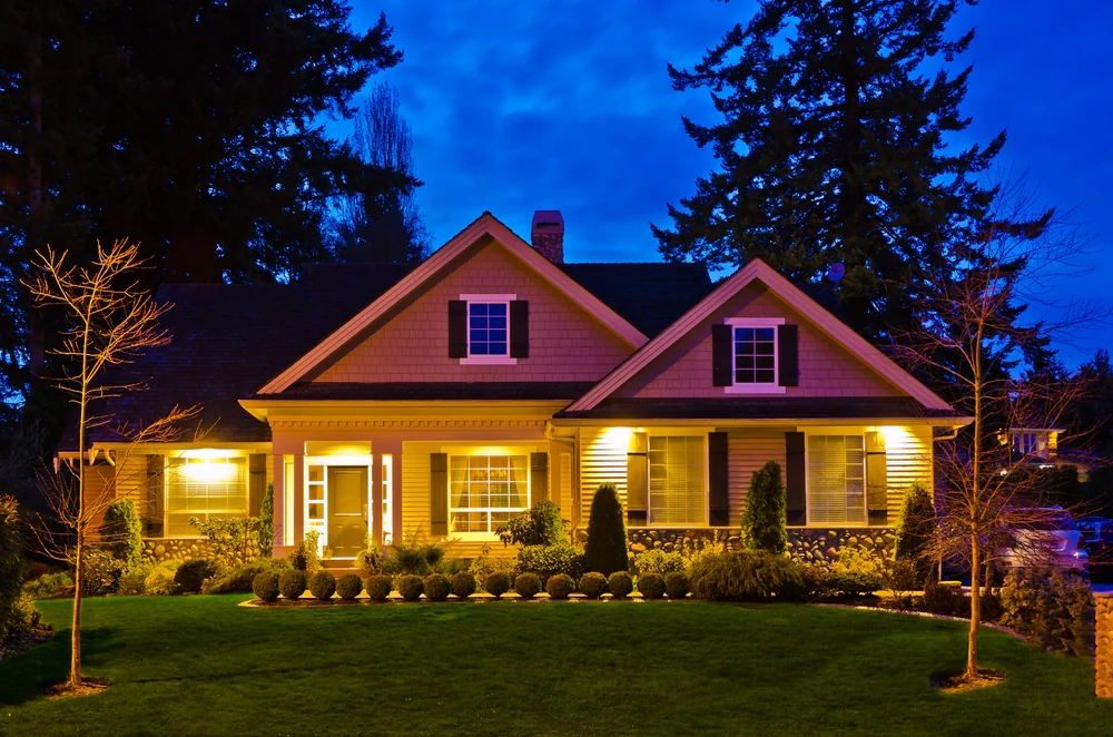 A cozy house with yellow lights glowing from the windows, surrounded by a neat lawn and small bushes, stands against a deep blue evening sky with tall trees in the background.