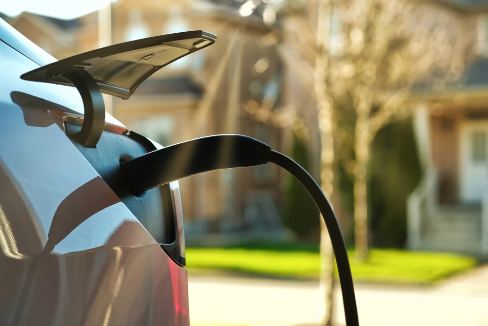 Close-up of an electric car being charged in a residential neighborhood, with the charging cable plugged into the vehicle and houses blurred in the background.
