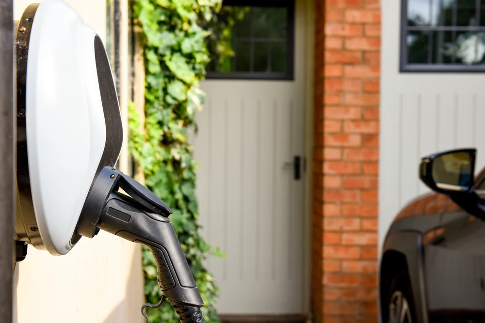 Close-up of an electric vehicle charging station mounted on an exterior wall near a brick house, with a black car parked nearby and green ivy growing beside the door.
