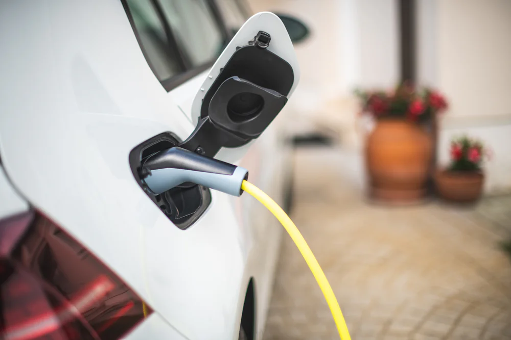 Close-up of an electric car being charged, with a yellow charging cable plugged into the vehicle. Blurred potted plants are visible in the background on a tiled outdoor area.