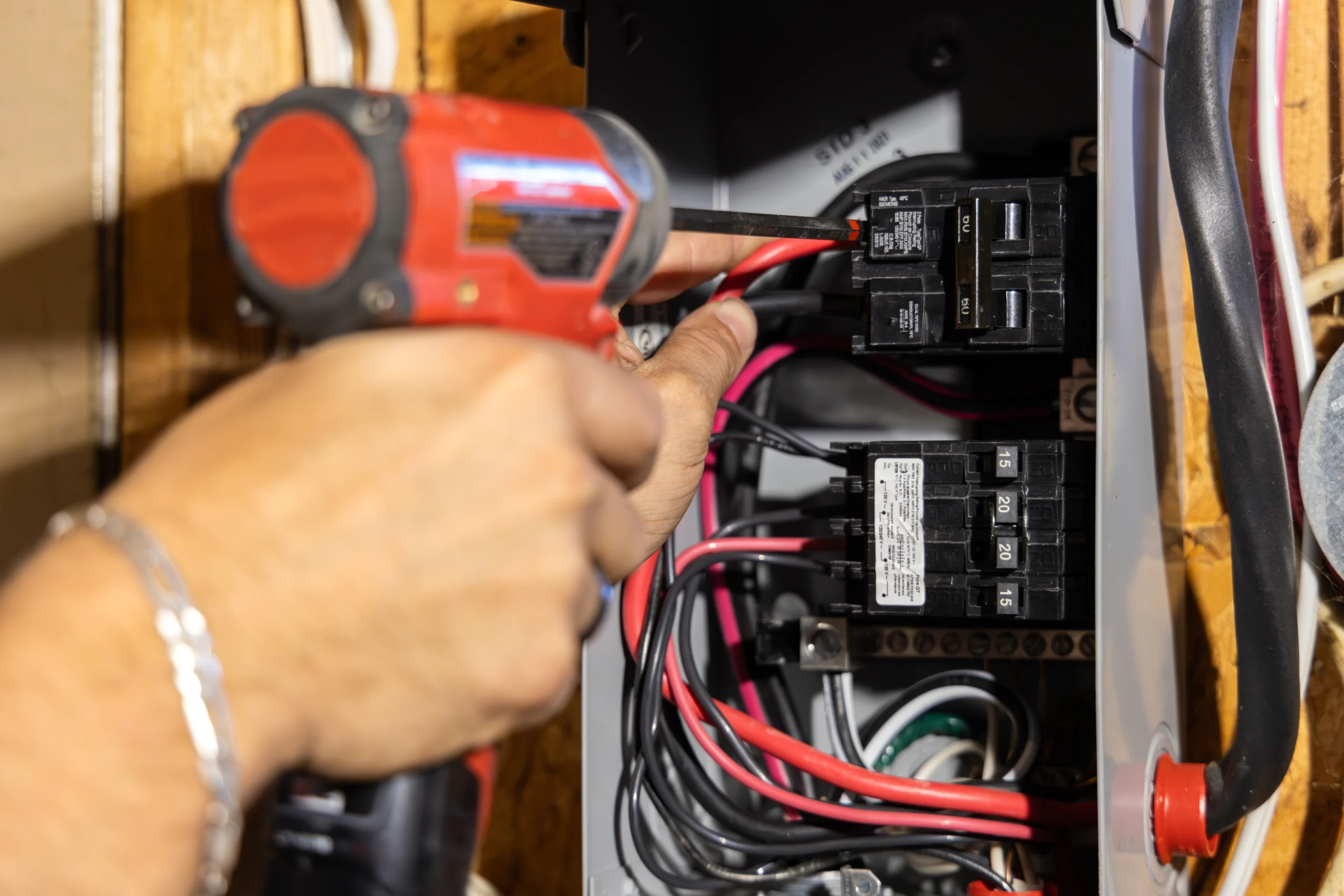 A person uses a red cordless drill to work on an open electrical panel with various wires and circuit breakers inside.