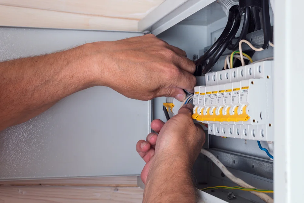 A person uses a screwdriver to adjust or repair switches inside an electrical panel, with several wires and yellow switches visible.