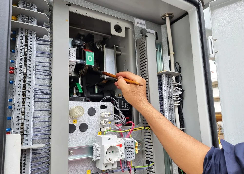 A person uses a brush to clean the interior components of an open electrical control panel, which contains wires, switches, and electrical parts.