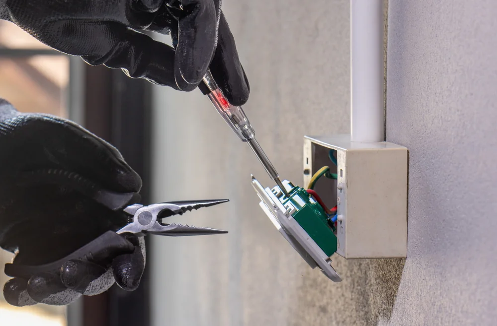 A person wearing black gloves uses a screwdriver and pliers to work on an exposed electrical outlet box mounted on a wall, with wires and internal components visible.