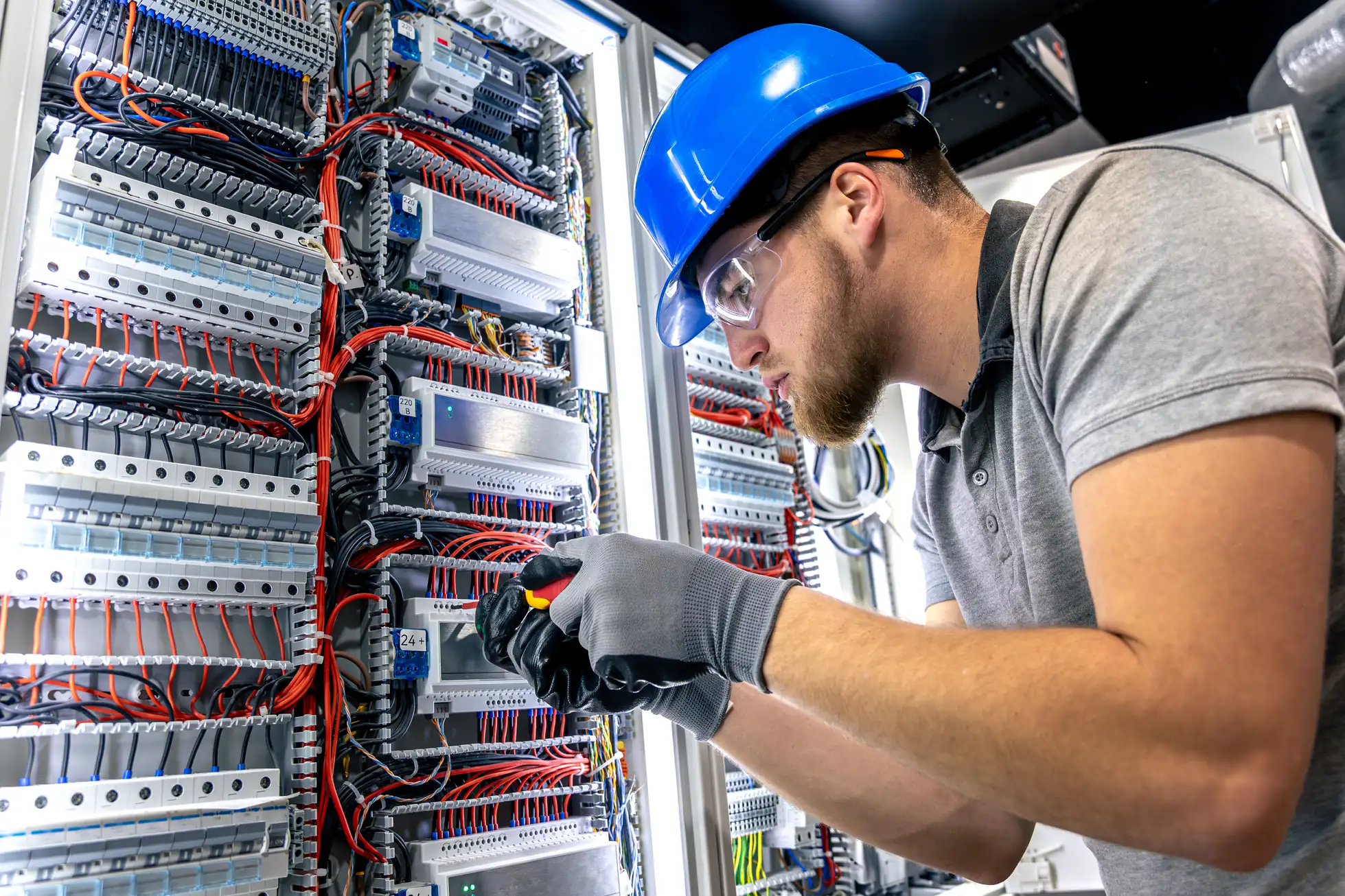 A technician wearing a blue hard hat, safety glasses, and gloves works on wiring inside an open electrical control panel filled with numerous cables, switches, and components.