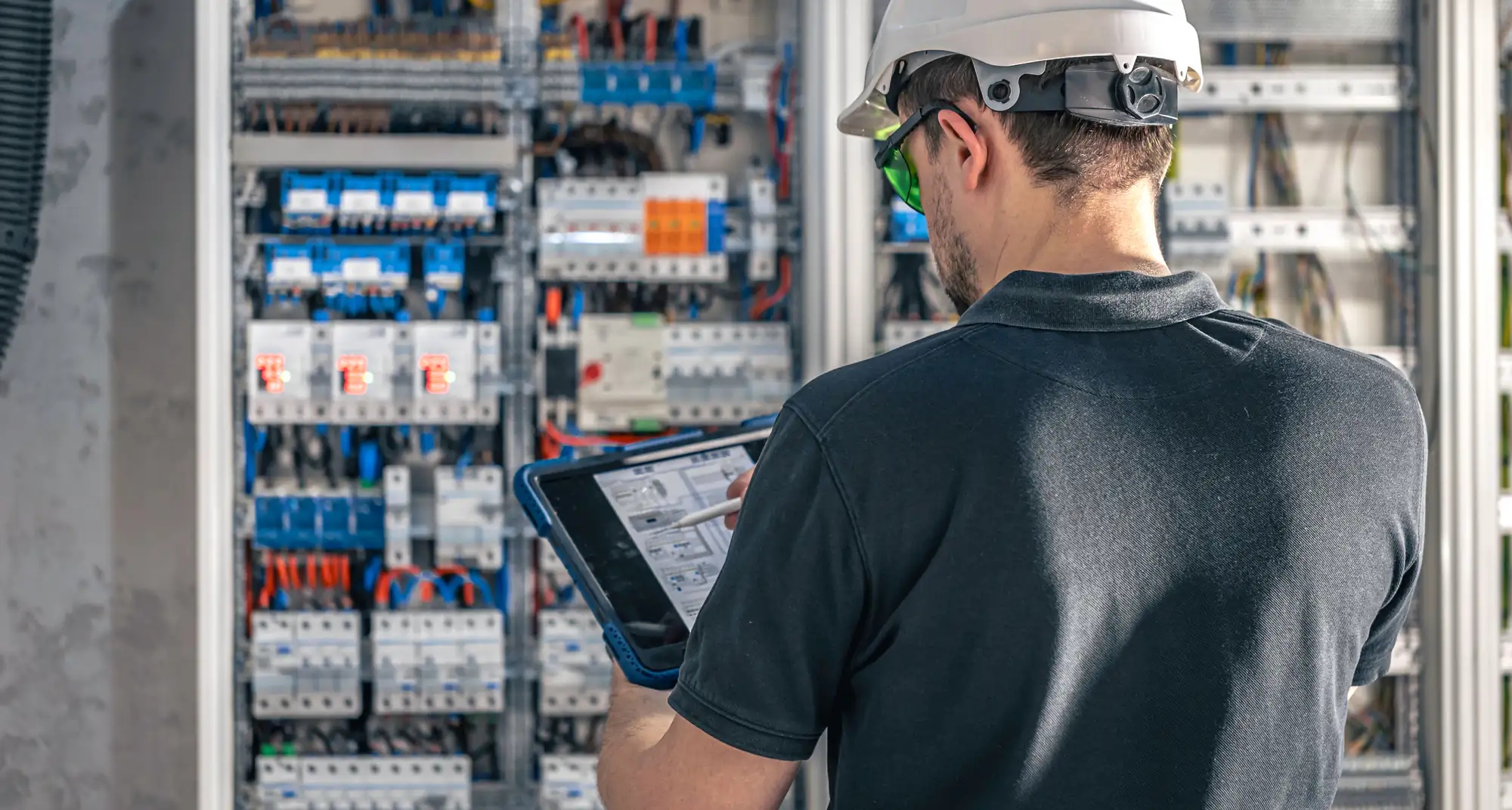 A technician wearing a white hard hat and safety glasses uses a tablet while inspecting an open electrical control panel filled with wires and components.