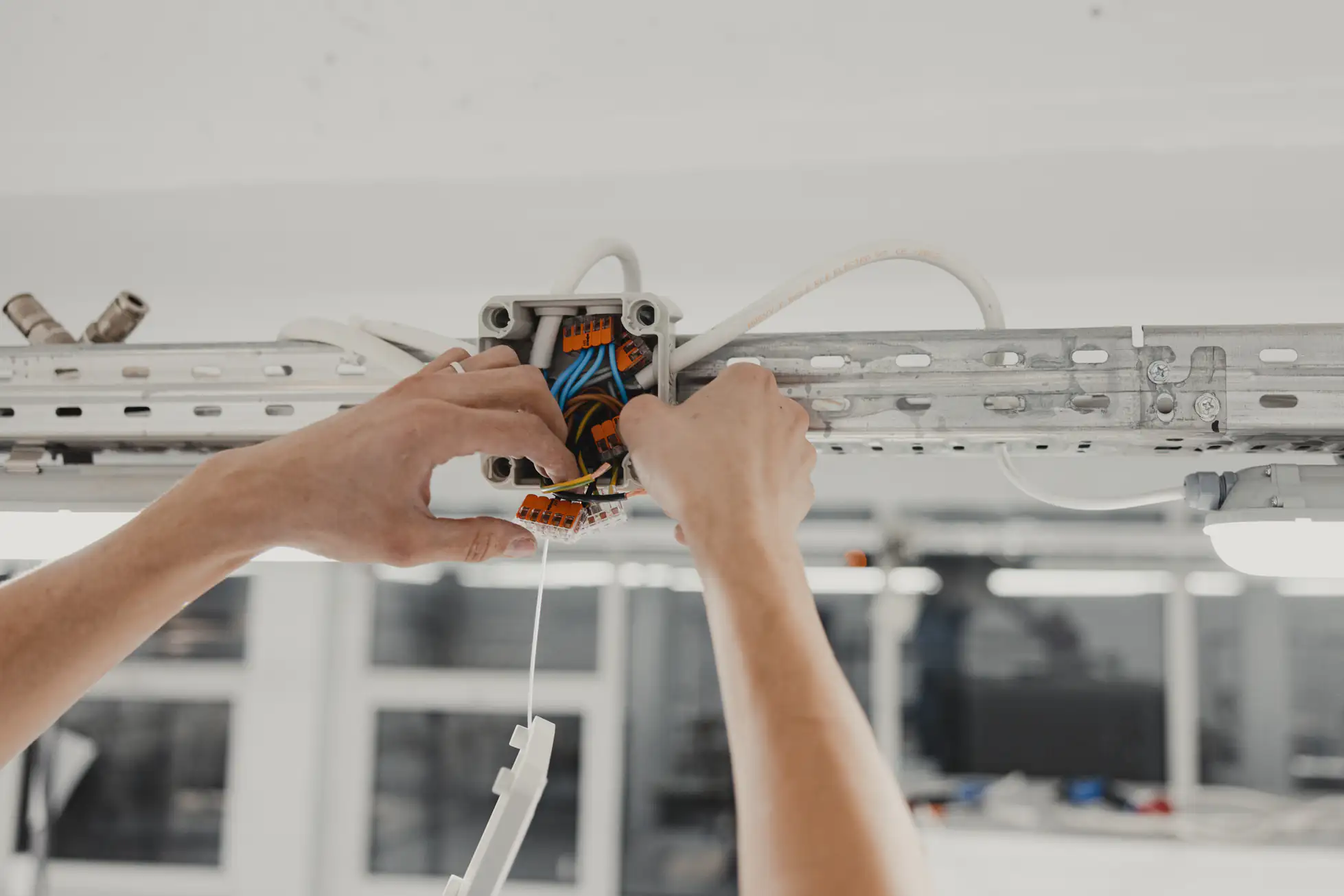 A person connects wires inside an electrical junction box mounted on a metal rail, with white cables and tools visible, in a well-lit indoor setting.