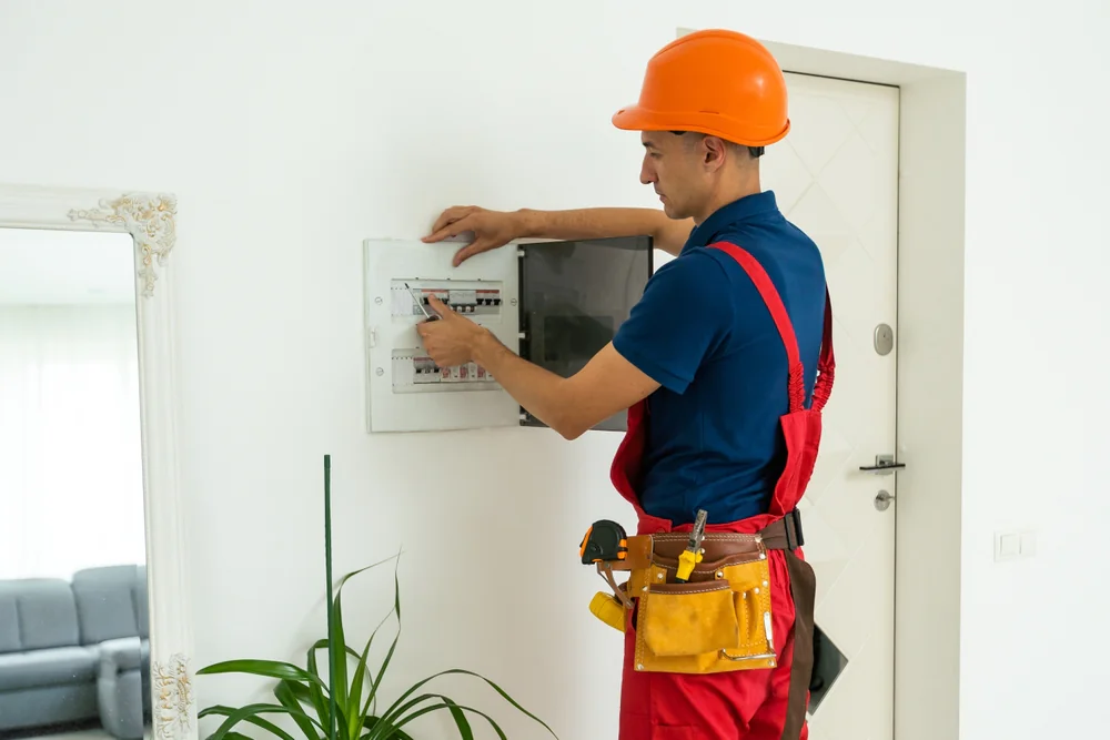 An electrician wearing an orange hard hat and red overalls uses tools while inspecting an open electrical panel on a white wall in a modern home. A plant and a mirror are visible nearby.