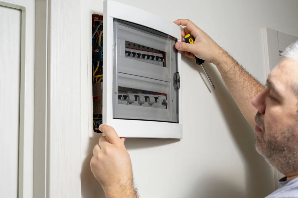 A person opens an electrical breaker panel on a wall with one hand while holding a screwdriver in the other hand, examining the switches and wiring inside.