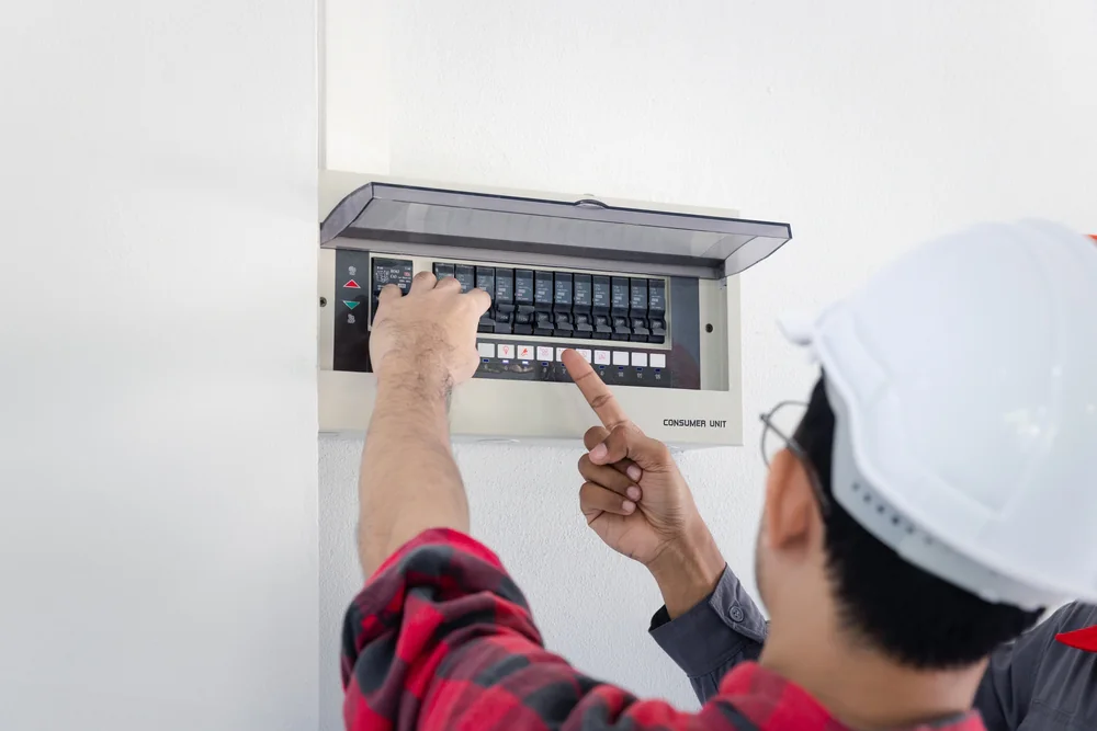 An electrician wearing a white safety helmet and plaid shirt points at and operates switches on an electrical breaker panel mounted on a white wall.