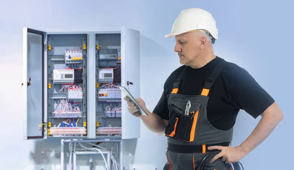 An electrician wearing a white hard hat and overalls stands in front of an open electrical control panel, looking at a tablet while holding cables, with tools visible in his pocket.