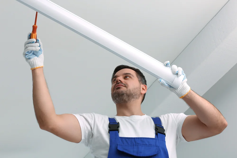 A man in work gloves and blue overalls uses a screwdriver to install or repair a fluorescent light fixture on a white ceiling.