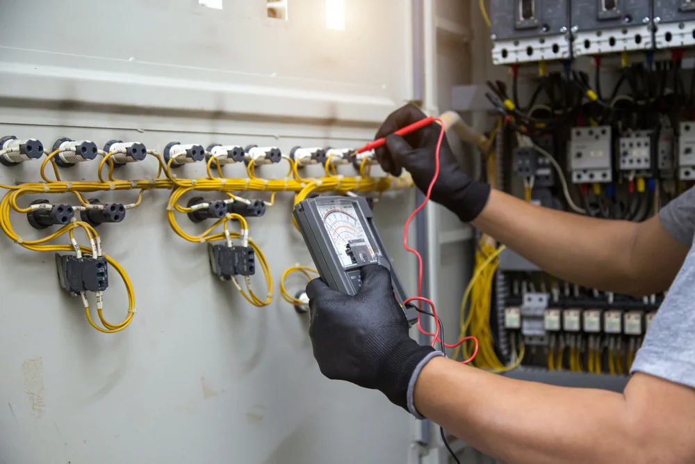 A person wearing black gloves uses a multimeter to check electrical wiring inside a control panel with yellow wires and various electrical components.