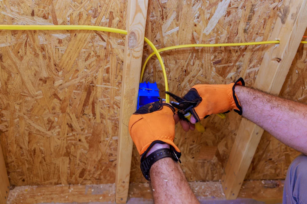 A person wearing orange gloves installs electrical wiring into a blue outlet box mounted on a wooden wall frame, using pliers.