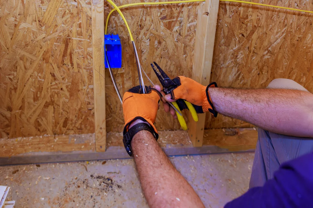 A person wearing orange gloves uses wire strippers to prep electrical wires for installation in a blue outlet box mounted on a wooden wall frame.