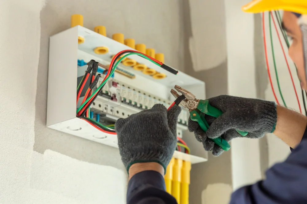 An electrician wearing gloves and a yellow helmet uses pliers to work on wiring inside an electrical panel mounted on a wall. Several colorful wires are visible connected to the panel.