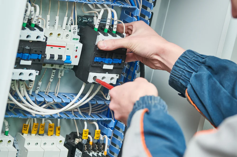 A person wearing a work uniform uses a screwdriver to adjust wiring in an electrical control panel filled with switches, relays, and cables.