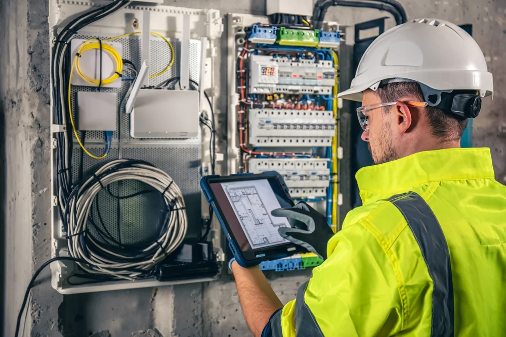 An electrician in a high-visibility jacket and hard hat inspects a wiring cabinet, holding a tablet displaying technical drawings, with cables and circuit components visible in an industrial setting.