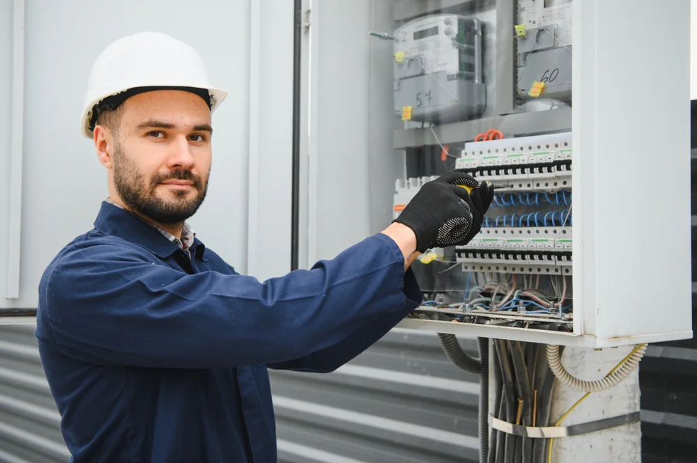 A man wearing a white hard hat, gloves, and blue coveralls adjusts wires inside an open electrical panel, looking at the camera, in an industrial setting.