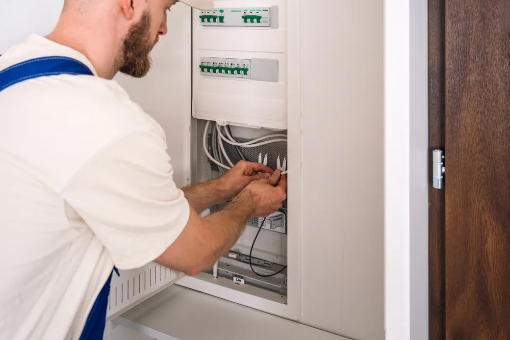 A person wearing a white shirt and blue overalls is working on an electrical panel, connecting or adjusting wires inside a wall-mounted cabinet.