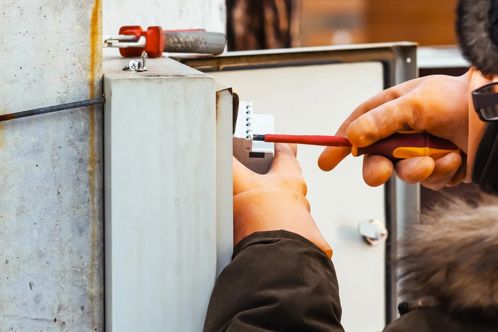 Person wearing protective gloves uses a screwdriver to work on electrical components inside an outdoor electrical panel or box, ensuring safety while performing maintenance or repairs.
