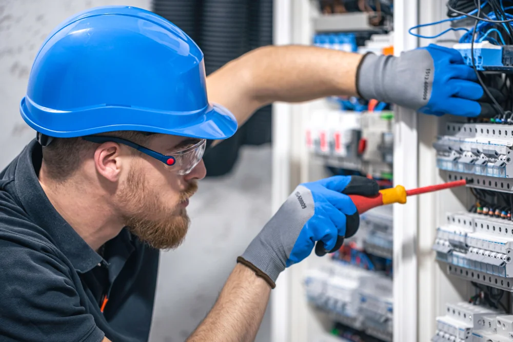 An electrician wearing a blue hard hat, safety glasses, and gloves uses a screwdriver to work on electrical panels with various wires and components.