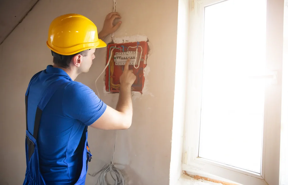A person wearing a yellow hard hat and blue shirt works on an electrical panel inside a building, using one hand to point at the wiring near a window with natural light coming in.