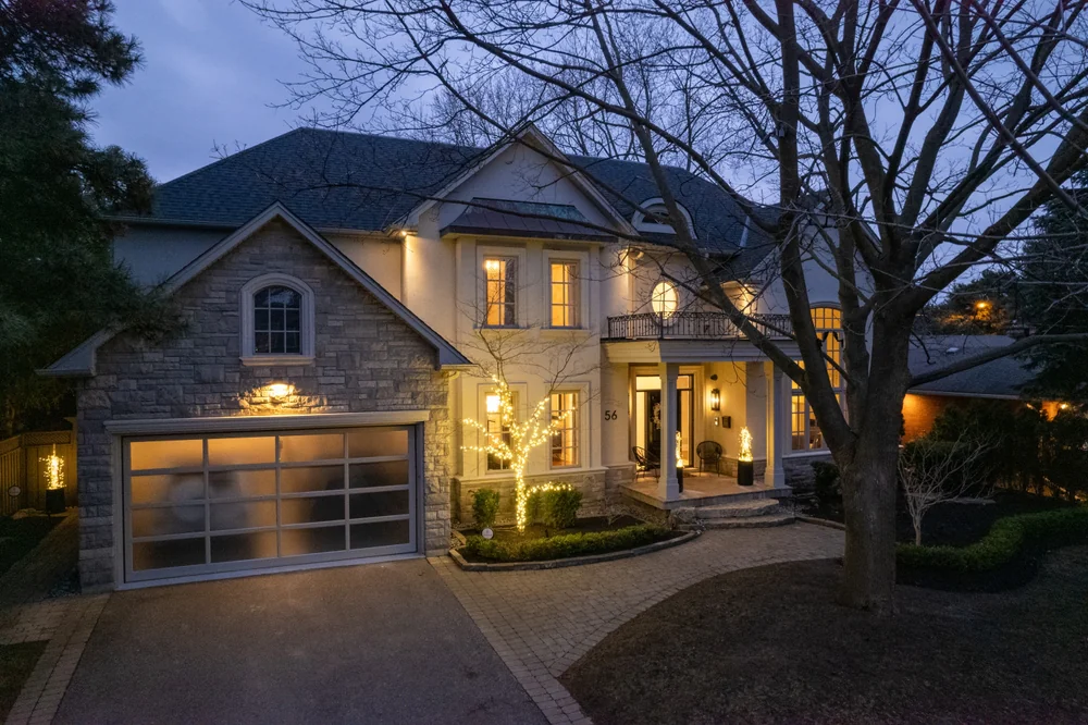 A large, two-story house with stone and stucco exterior is lit warmly at dusk. Trees frame the driveway, and soft exterior lights highlight the entrance and garage.