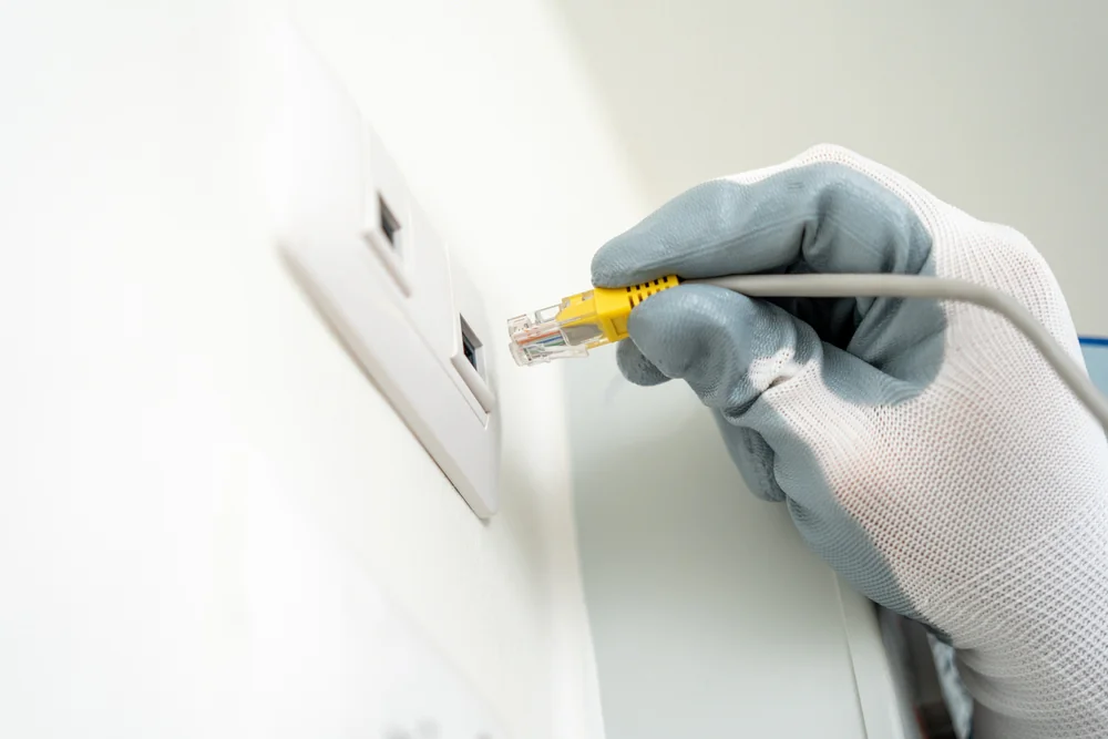 A person wearing white and gray gloves is plugging a yellow Ethernet cable into a wall network socket.