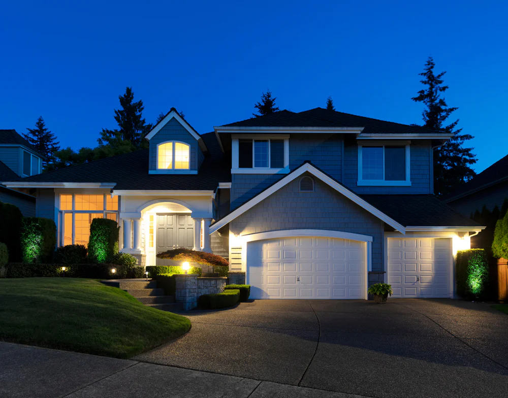 A two-story suburban house with blue-gray siding and white trim is lit up at dusk. The driveway leads to a double garage, and the front yard is landscaped with grass and shrubs. Lights glow from the windows and entryway.