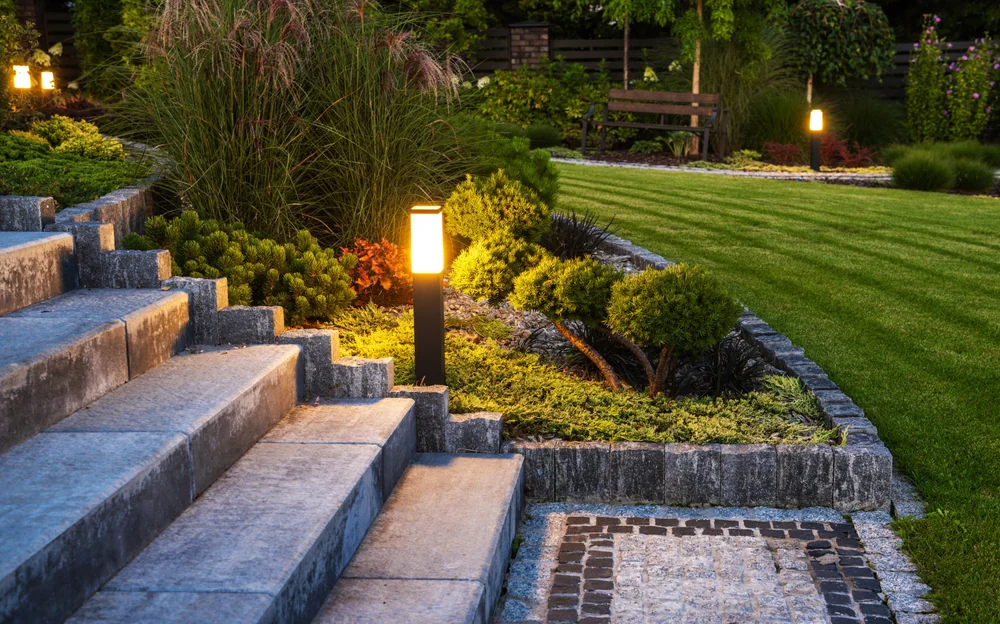 Stone steps lead to a landscaped garden with glowing pathway lights, neatly trimmed grass, shrubs, and ornamental plants. A wooden bench sits in the background near a dark fence.